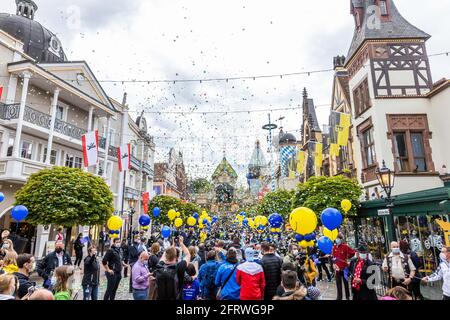 Ruggine, Germania. 21 Maggio 2021. I visitatori di Europa-Park camminano attraverso un viale all'ingresso del parco mentre i dipendenti del parco si levano in piedi guardia e confetti piovono giù dai cannoni confetti. Europa-Park a Baden-Württemberg è uno dei primi parchi a tema più importanti della Germania ad aprire le porte ai visitatori. Il parco di Rust ha accolto nuovamente i visitatori dopo mesi di pausa forzata Corona - come parte di un progetto modello approvato dallo stato. Credit: Philippe von Ditfurth/dpa/Alamy Live News Foto Stock