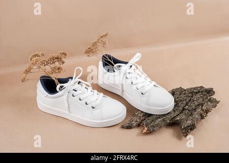 Concetto etico di scarpe vegan. Un paio di sneakers bianche con fiori secchi su corteccia di albero, sfondo neutro di carta da artigianato beige. Foto Stock