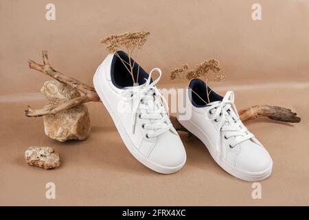 Concetto etico di scarpe vegan. Un paio di sneakers bianche con fiori secchi sul legno e mucchio di pietre, fondo carta da artigianato beige neutro. Foto Stock