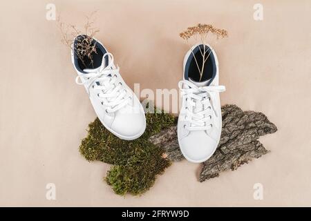 Concetto etico di scarpe vegan. Un paio di sneakers bianche con fiori secchi su corteccia di alberi e muschio, fondo di carta da mestiere beige neutro. Vista dall'alto, disposizione piatta. Foto Stock