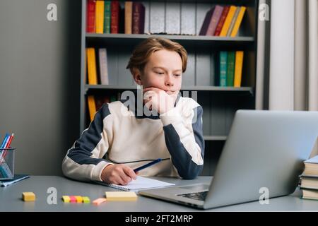 Stanco ragazzo annoiato guardando lo schermo del notebook durante la lezione online, preparandosi a prendere appunti nel notebook. Foto Stock