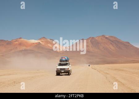 Sullo sfondo di vulcani, un veicolo fuoristrada 4x4 attraversa il paesaggio desertico della Bolivia in un safari turistico terrestre. Foto Stock