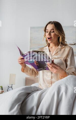 Donna entusiasta di leggere rivista mentre si siede a letto la mattina Foto Stock