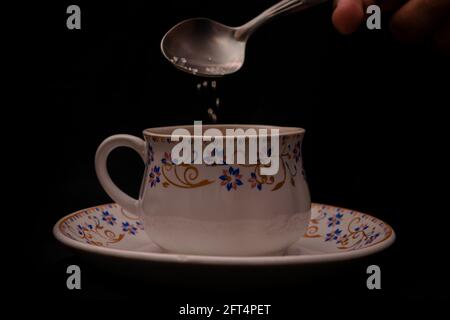 Mano aggiungendo un cucchiaio di zucchero ad una tazza di Tè indiano masala su sfondo nero Foto Stock