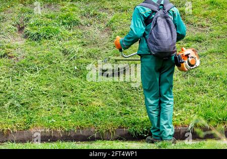 Un operaio in tute verdi e uno zaino sulla schiena falda l'erba con un trimmer industriale a benzina in un parco cittadino. Spazio di copia. Foto Stock