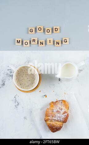 Buona colazione del mattino di concetto minimo. Tazza di caffè, croissant e testo Buongiorno su sfondo grigio rosa. Disposizione piatta Foto Stock