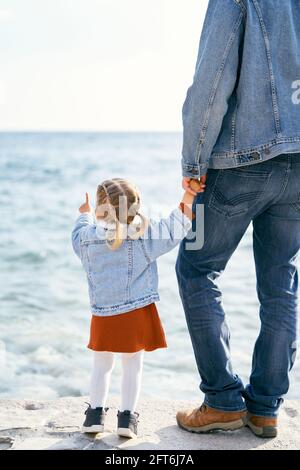 Papà in una giacca in denim e jeans tiene la mano di una bambina che punta al mare, in piedi sulle pietre vicino all'acqua. Vista posteriore Foto Stock