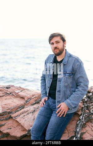 Uomo pensivo in jeans e giacca in denim in posa seduta su pietre sullo sfondo del mare. Vista laterale Foto Stock