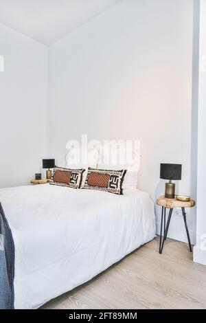 Interni dallo stile minimalista di una camera da letto bianca e confortevoli letto decorato con cuscini ornamentali posti ad angolo e con lampade sui comodini Foto Stock