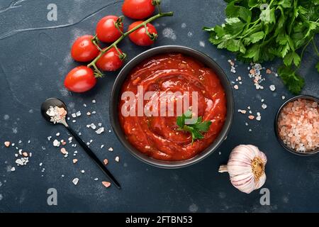 Salsa piccante al ketchup di pomodoro con ingredienti prezzemolo, aglio e pepe in un recipiente nero con spezie e pomodori freschi su un'ardesia scura, pietra o concre Foto Stock