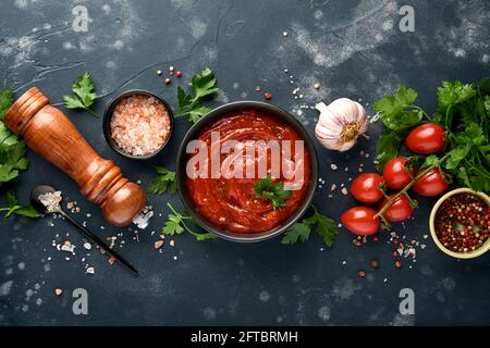 Salsa piccante al ketchup di pomodoro con ingredienti prezzemolo, aglio e pepe in un recipiente nero con spezie e pomodori freschi su un'ardesia scura, pietra o concre Foto Stock