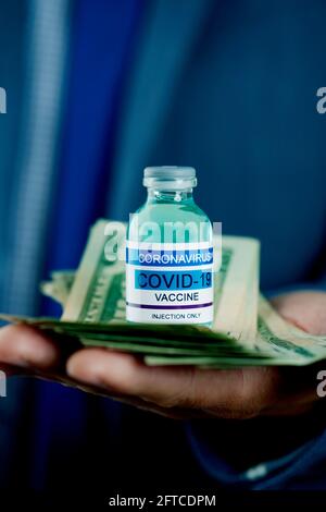 closeup di un uomo d'affari, indossando un vestito blu, con una bottiglia simulata di vaccino covid-19 e un mazzo di banconote in dollari nelle sue mani Foto Stock
