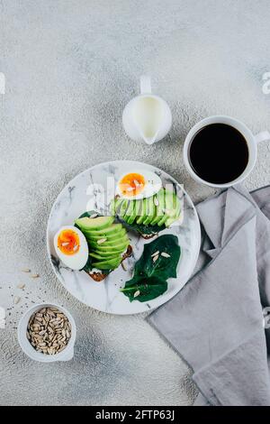 Delizioso toast su pane a grani di segale scuro preparato con avocado fresco affettato, formaggio cremoso, spinaci e semi. Tazza di caffè nero. Colazione salutare Concep Foto Stock