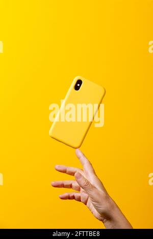 Mano donna matura che tiene uno smartphone giallo, con una custodia gialla, con un dito, su uno sfondo giallo Foto Stock