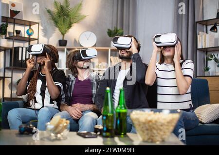 Giovani multirazziali che si rilassano insieme sul divano e giocano a videogame in occhiali VR. Felici gli amici che usano la tecnologia moderna mentre trascorrono il tempo a casa. Foto Stock