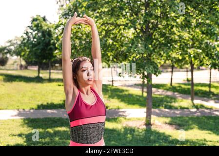 presa orizzontale di una donna caucasica che allunga le braccia su un parco. Indossa abiti sportivi rosa e grigi Foto Stock
