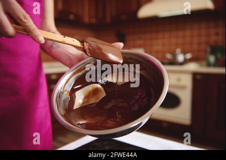 Pasticciere produttore di pasticceria al cioccolato mescolando la massa di cioccolato fuso in una ciotola d'acciaio con un cucchiaio di legno. Produzione di cioccolato fatto in casa Foto Stock