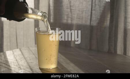 l'uomo versa la birra allo zenzero dalla bottiglia di vetro in bicchiere sul tavolo di quercia bianca con luce del tramonto, foto ampia Foto Stock