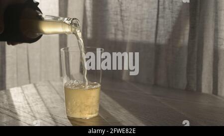 l'uomo versa la birra allo zenzero dalla bottiglia di vetro in bicchiere sul tavolo di quercia bianca con luce del tramonto, foto ampia Foto Stock