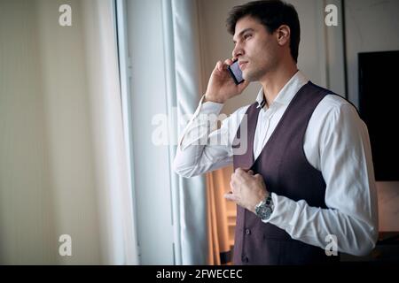 premuroso giovane caucasico che parla su un cellulare vicino alla finestra, fissando i bottoni sul suo giubbotto con un'altra mano Foto Stock
