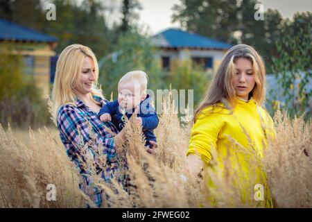 Una giovane madre cammina con i suoi figli in un campo con erba alta. La mamma tiene il ragazzo tra le braccia. Una famiglia felice Foto Stock