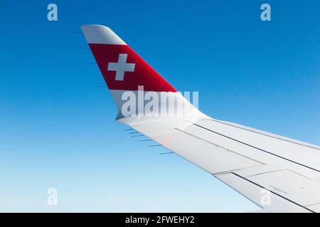 Ali dell'aereo che mostrano la bandiera svizzera / ala di un aereo della serie Bombardier C e la vista di cielo azzurro soleggiato a crociera in altitudine in una giornata limpida con cielo blu. (100) Foto Stock