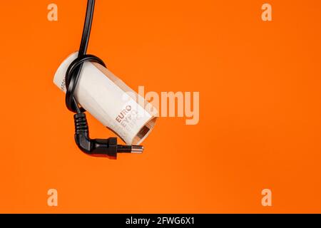 Le banconote in euro sono legate in un nodo da un cavo elettrico nero con una spina su uno sfondo arancione colorato. Primo piano. Concetto creativo di risparmio di energia elettrica c Foto Stock