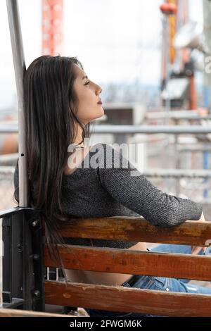 giovane donna con lunghi capelli neri seduta su una panca di legno con il viso in profilo, riposato in un parco all'aperto, stile di vita e bellezza Foto Stock