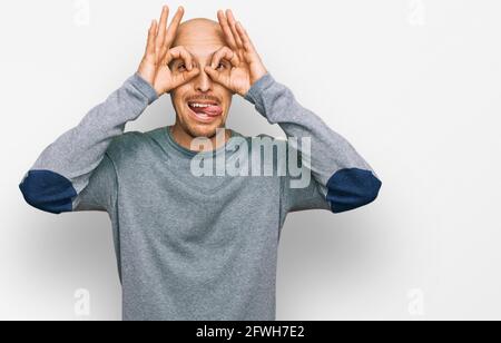 Uomo calvo con barba che indossa abiti casual facendo un gesto ok come binocolo che si stacca la lingua, occhi che guardano attraverso le dita. Espressione pazza. Foto Stock
