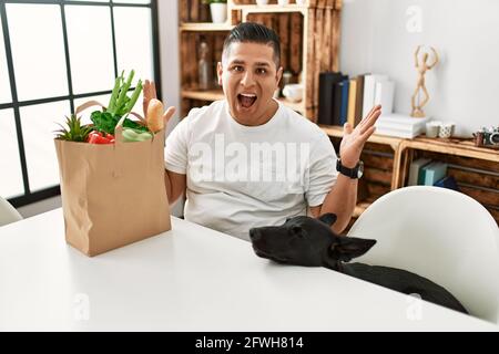 Giovane uomo ispanico seduto con borsa di carta con generi alimentari festeggiando vittoria con sorriso felice e espressione vincente con mani alzate Foto Stock