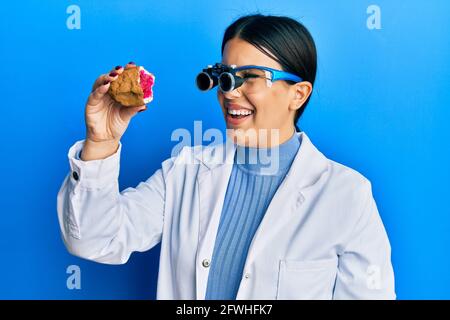 Bella donna gioielliera bruna che tiene geode pietra indossare occhiali di ingrandimento sorridente con un sorriso felice e fresco sul viso. Mostrando i denti. Foto Stock