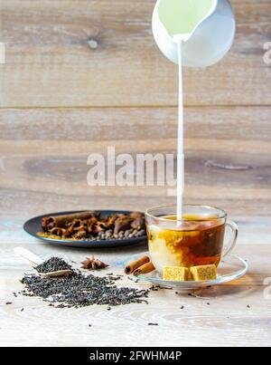 Bevanda indiana tradizionale - tè masala con spezie sfondo di legno. Versate il latte dal contenitore del latte in una tazza di vetro con il tè. Foto Stock