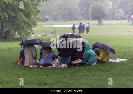 Gruppo riparato sotto gli ombrelloni durante un improvviso temporale in un parco. Persone catturate dalla pioggia durante il downpour, durante la pandemia COVID 19 Foto Stock