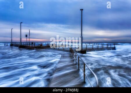 Spiaggia di Mona vale piscina rocciosa ad alta marea tagliata da onde alte durante l'ora di alba blu con nuotatori attivi. Foto Stock