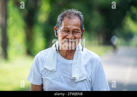 Ritratto dell'uomo anziano asiatico in abbigliamento fitness che corre in un parco. Primo piano di un uomo sorridente che corre mentre ascolta la musica con gli auricolari. El sano Foto Stock