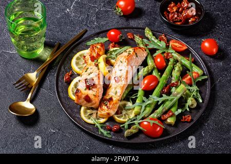 salmone al forno con limone e pancetta caramellate servito con asparagi insalata di rucola selvatica di pomodori ciliegini su un piatto nero con posate dorate Foto Stock