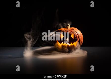 Halloween, zucca arancione con un viso spaventoso e luminoso su uno sfondo scuro. Esce fumo grigio spesso. Primo piano di jack-o'-lanterna alla vigilia di tutti Foto Stock