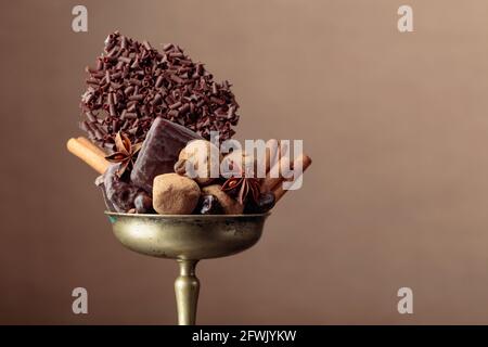 Vari cioccolatini con ingredienti. In una vecchia ciotola di ottone caramella, chicchi di caffè, cannella, anice e pezzi di cioccolato nero rotto. Foto Stock