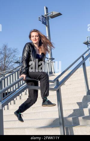 giovane donna in giacca di pelle seduta all'aperto su una scala ringhiera Foto Stock