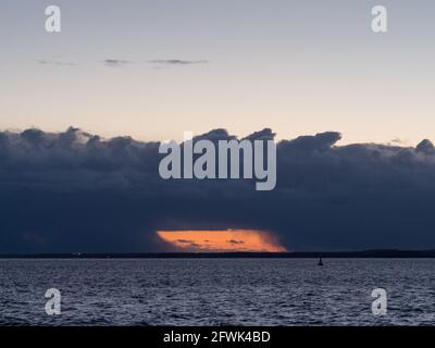 Una luce arancione brillante che appare come una finestra attraverso Nubi minacciose sopra le acque del mare solente Foto Stock