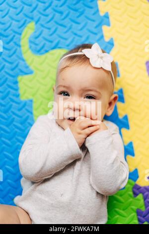 Piccola bambina con un fiore sulla testa dentro una blusa bianca giace su un tappeto multicolore con lei mani in bocca Foto Stock
