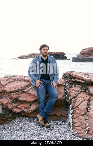 Uomo pusivo in jeans e giacca in denim in posa in piedi la riva pendente su pietre sullo sfondo del mare Foto Stock