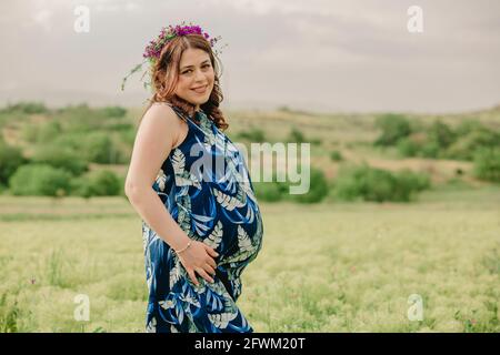 Gravidanza e maternità. Giovane donna incinta con fiori selvatici sulla testa in piedi in campo, tenendo il suo grande ventre e in posa per la macchina fotografica Foto Stock