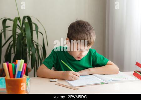 bambino di 7 anni che fa lezioni seduto alla scrivania nella sua stanza. I bambini scrivono i compiti in un notebook Foto Stock