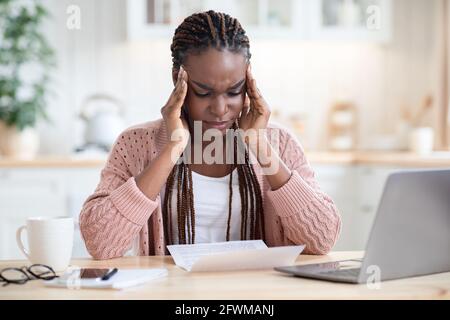 Stressata donna nera che legge documenti mentre si siede al tavolo dentro Cucina Foto Stock