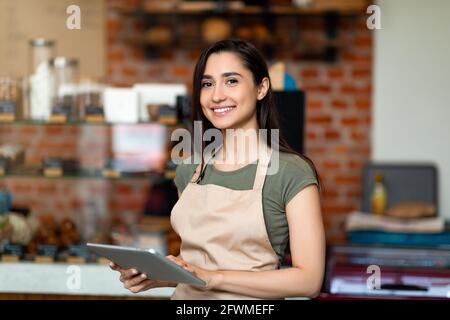 Aprire una piccola impresa. Donna araba felice in grembiule vicino al bancone della barra che tiene il tablet digitale e guarda la fotocamera Foto Stock