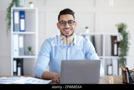 Felice giovane uomo d'affari arabo in occhiali seduti alla scrivania, lavorando su un computer portatile presso l'ufficio aziendale Foto Stock