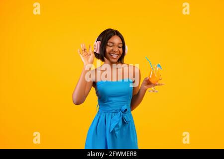 Donna nera in outfit elegante che tiene cocktail esotici e godere la musica preferita nelle cuffie su sfondo arancione Foto Stock