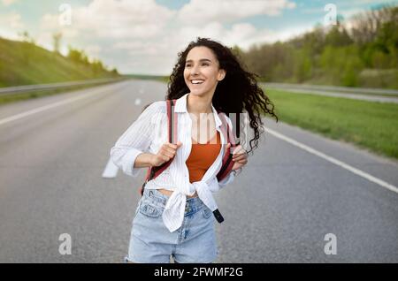 Bella giovane donna con zaino a piedi lungo l'autostrada, viaggiando in autosstop, hitchtrekking in campagna Foto Stock