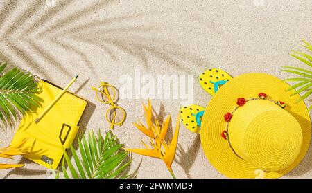 Cappello rotondo giallo, blocco note e occhiali da sole con fiori tropicali arancio e foglia di palma sulla spiaggia di sabbia. Foto Stock